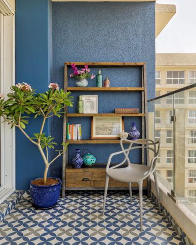 Balcony with textured blue wall, wooden shelf display, patterned tile floor, and potted frangipani plant.