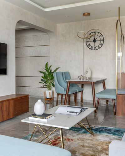 Living and dining area with teal velvet chairs, marble coffee table, wall clock, and arched niche.