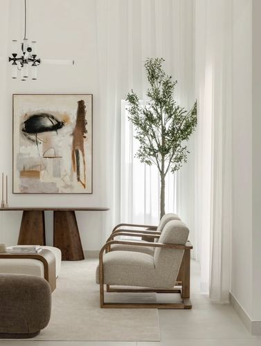Neutral living room with armchairs, carved wood console, abstract art, and sheer curtains framing an indoor tree