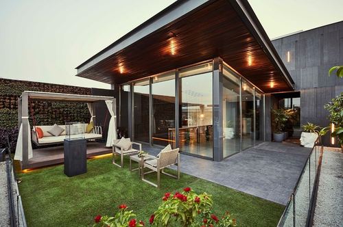 Rooftop terrace with cabana daybed, vertical garden, and glass-walled room under wood-slat canopy at dusk