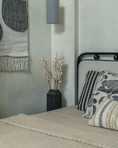 Bedroom corner with black iron headboard, textured cushions, dried grass in ceramic vase, striped pendant lamp