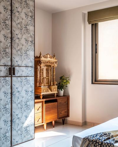 Bedroom corner with floral-paneled wardrobe, carved wooden mandir on dresser, sunlit window