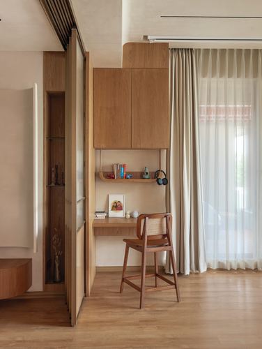 Wooden study nook with floating shelf, cabinet, and cane-backed chair beside sheer curtains