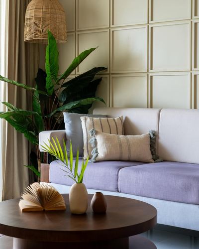 Living room corner with lavender sofa, paneled wall, rattan pendant, and round wooden coffee table with vases