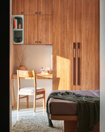 Sunlit bedroom corner with wooden wardrobe, built-in study desk, chair, and bed with green throw