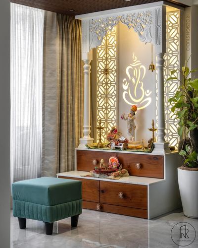 Backlit pooja unit with carved white arch, jali panels, wooden drawers, and a green velvet stool beside sheer curtains