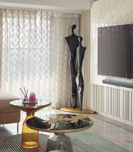 Living room corner with abstract black sculptures, nested glass side tables, and sheer patterned curtains beside a marble tv wall