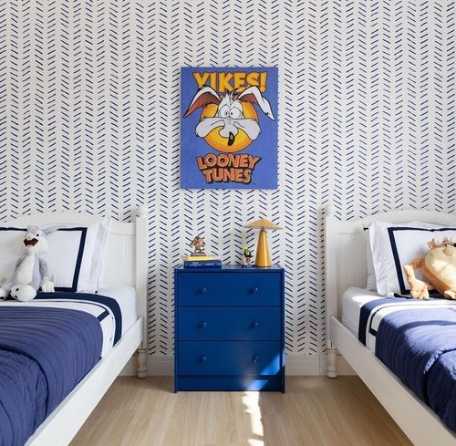 Kids bedroom with two white twin beds, navy bedding, blue dresser, and Looney Tunes poster on herringbone wallpaper