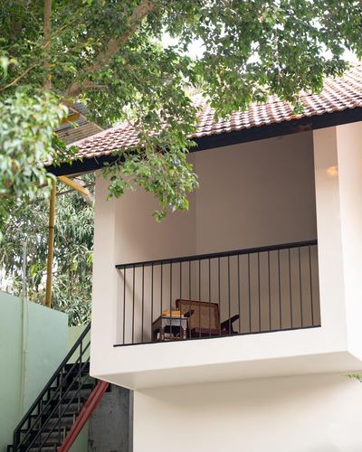 Exterior view of a shaded upper-floor balcony with cane chair, framed by tree canopy and tiled roof