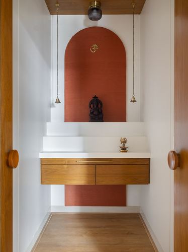 Recessed pooja niche with arched terracotta panel, hanging brass bells, and floating wood cabinet.