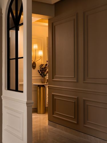 View through a paneled doorway into a warmly lit hallway with a gold pedestal console table and wall sconces