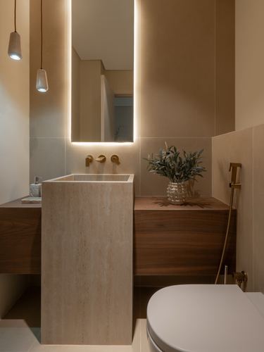 Powder room with travertine pedestal sink, backlit mirror, and walnut vanity in warm tones