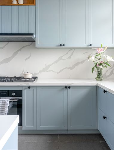 Corner of a pale blue shaker kitchen with white veined stone backsplash and countertop, flowers on counter