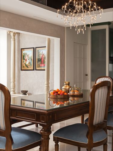 Wooden dining table with striped chairs beneath chandelier, framed miniatures and carved columns in niche behind