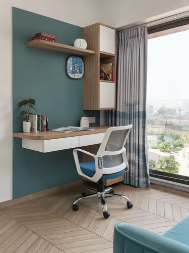 Study nook with floating wood desk, teal wall, wall cabinet, and mesh office chair by window