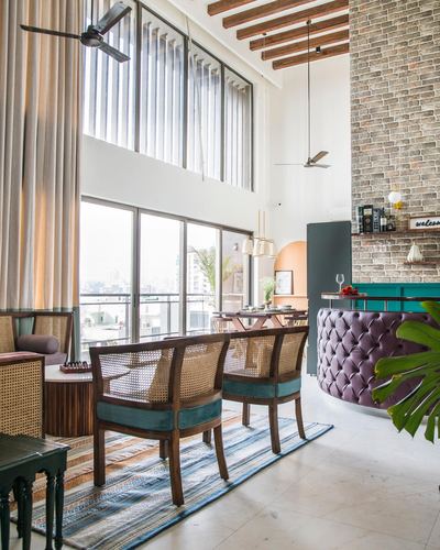 Double-height living room with cane chairs, exposed brick wall, and a tufted mauve bar counter