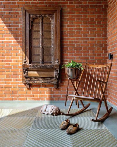 Exposed brick porch corner with carved wooden window frame, rocking chair, plant stand, and sleeping dog on patterned tile floor