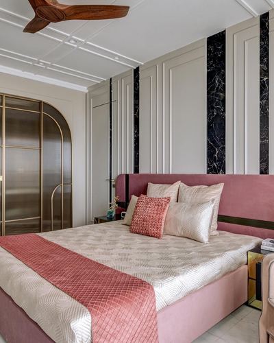 Pink upholstered bed against beige panelled wall with black marble strips and arched brass wardrobe.