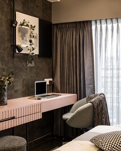 Pink fluted study desk with laptop against dark tiled wall, curtained window and grey chair beside bed