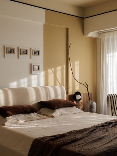Sunlit bedroom with mustard accent wall, cream upholstered headboard, brown linens and gallery photo frames