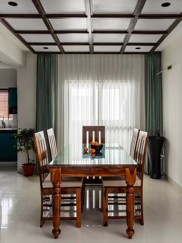 Wooden dining table with glass top and six chairs beneath a coffered ceiling, sage curtains behind