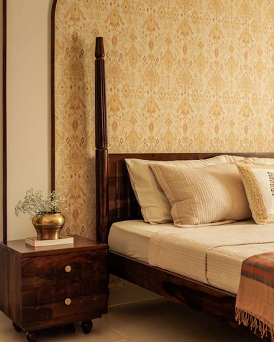 Four-poster wooden bed with cream linens against patterned gold wallpaper, brass planter on bedside table