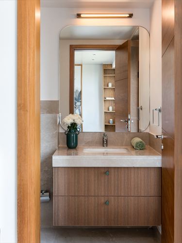 Bathroom vanity with fluted wood drawers, stone countertop, rounded mirror, and flower vase