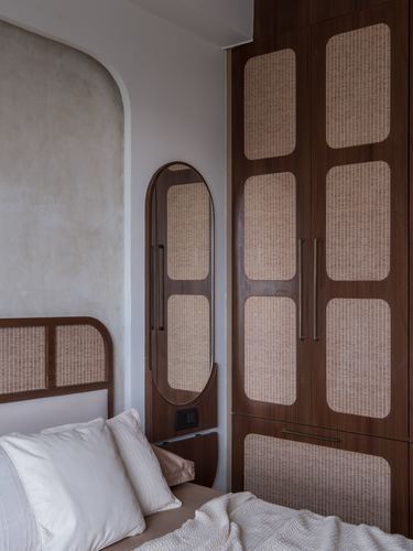 Wooden wardrobe with cane inlay panels beside an oval mirror in a bedroom with arched niche wall