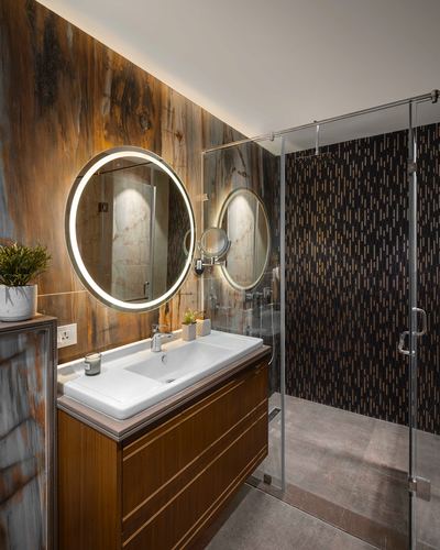 Bathroom with veined stone wall, backlit round mirror, wood vanity, and dark patterned shower tile