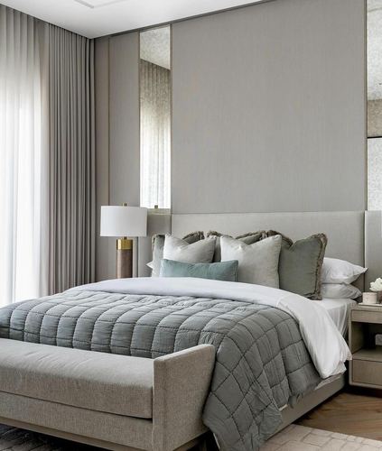 Grey upholstered bedroom with tall panelled headboard, bench, and layered green-grey pillows near sheer curtains