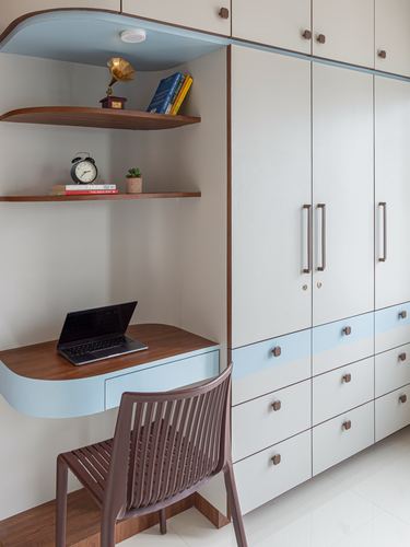 Curved wooden study desk with laptop beside a tall white wardrobe with brass handles