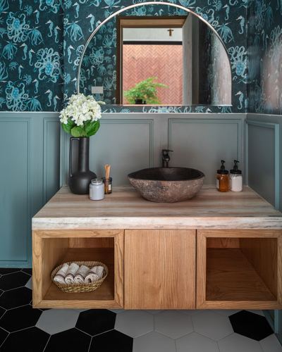 Bathroom vanity with stone vessel sink, wood cabinetry, arched mirror and patterned teal wallpaper
