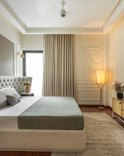 Beige bedroom with tufted headboard, sage throw, paneled walls, and glowing floor lamp beside curtained window