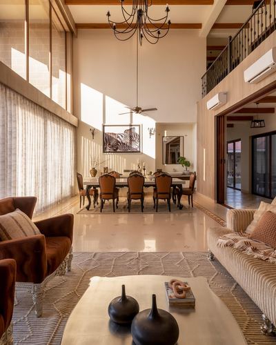 Double-height living and dining space with chandelier, carved chairs, sunlight through tall windows