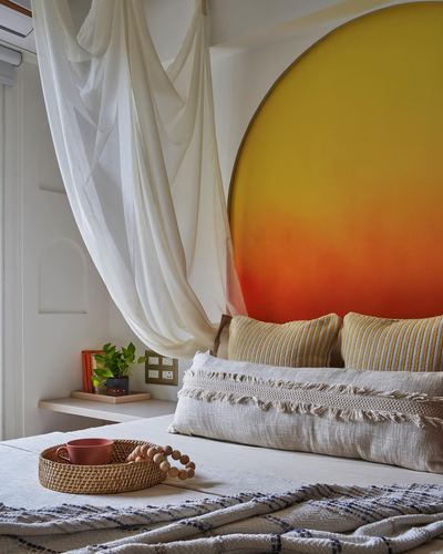 Bed with linen pillows below a circular orange-yellow sunset mural and draped white canopy fabric