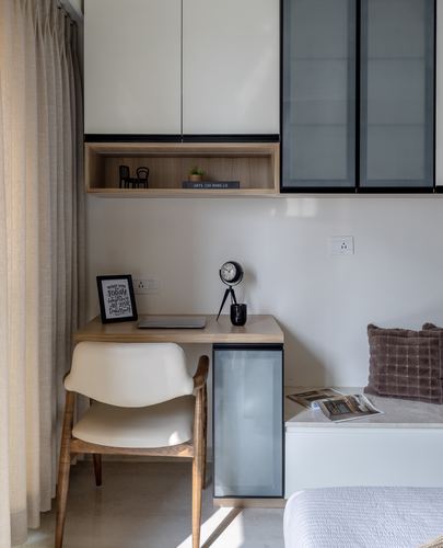 Small study desk with chair beneath overhead wood and glass cabinets near a window seat