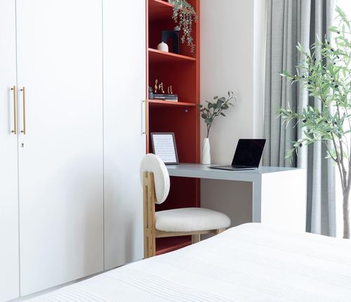 White wardrobe wall with red open shelving nook and built-in desk beside bed, daylight from right