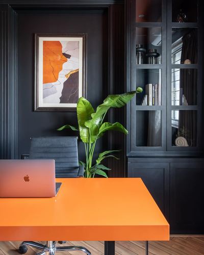 Home office with glossy orange desk against black paneled wall and glass cabinet