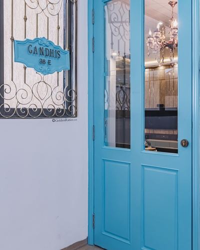 Turquoise glass-paned door beside white wall with ornate iron grille window and Gandhis nameplate.