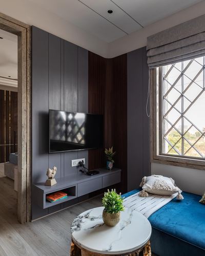 Living room corner with grey panelled TV wall, wood slats, marble table and blue velvet bench by a lattice window