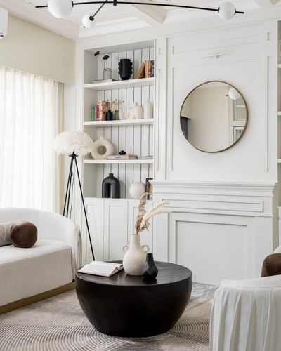 White paneled living room corner with round brass mirror, built-in shelves, and black round coffee table