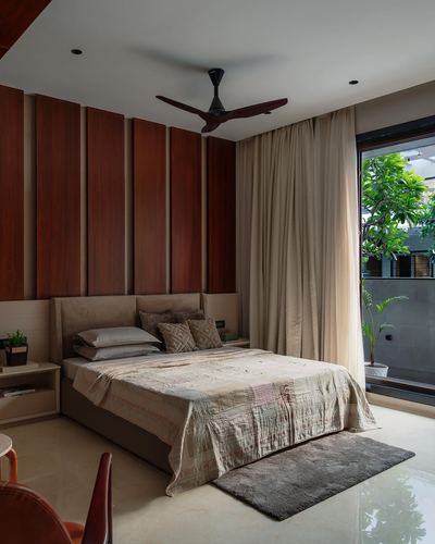 Bedroom with wood-panelled wall, upholstered bed, sheer curtains and glass door to greenery