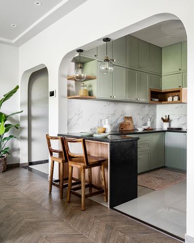 Sage green kitchen seen through an arched opening with cane barstools at a black-topped island