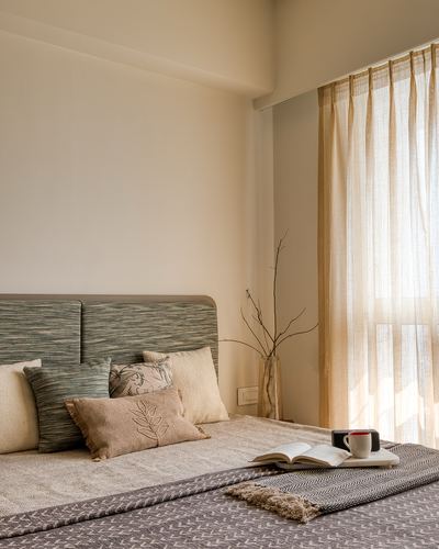 Bed with sage upholstered headboard, layered pillows, and sunlit sheer curtains beside a vase of branches