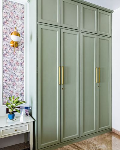 Sage green paneled wardrobe with brass handles beside a floral wallpaper niche and brass sconce