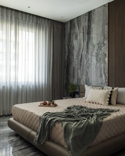 Bedroom with grey stone feature wall behind a quilted bed and sheer curtains