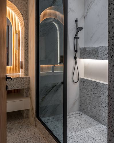Bathroom with terrazzo shower, black fixtures, and glowing arched marble niche beyond glass partition