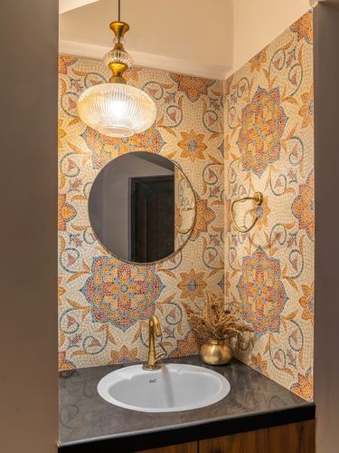 Powder room niche with floral mosaic tile walls, round brass-framed mirror, and dark stone counter with oval sink