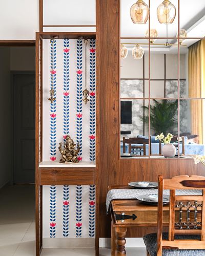 Wooden pooja unit with brass Ganesha idol against floral wallpaper, dining area reflected in glass beside it