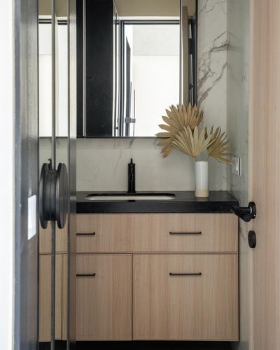Bathroom vanity with black countertop, wood cabinetry, marble backsplash, and dried palm arrangement